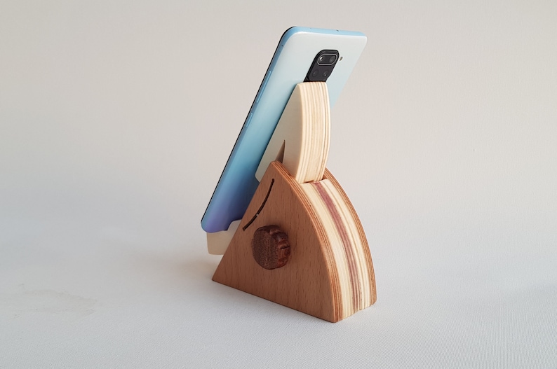 Op de afbeelding: Een houten telefoonstandaard in de vorm van een driehoek met een lichtblauwe telefoon erop. De standaard is gemaakt van licht en donker hout.
