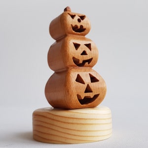 May include: A wooden Halloween decoration featuring three carved jack-o'-lanterns stacked on top of each other. The pumpkins have carved faces and are made of light brown wood. The base is a light-colored wooden cylinder.