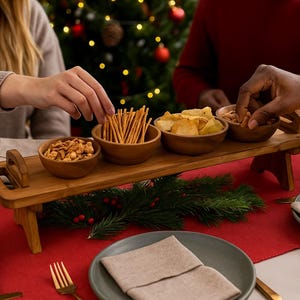 May include: A wooden serving tray with four small wooden bowls filled with snacks. The tray is on a red tablecloth, with plates, cutlery, and a lit candle. A Christmas tree is in the background.