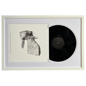 May include: A white framed print of a black and white illustration of a person's head with a blurred background. The print is next to a black vinyl record in a white frame.