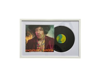 May include: A white framed picture with a black vinyl record and a photo of Jimi Hendrix. The record label says "Experience Hendrix" and the photo is from "The Best of Jimi Hendrix".