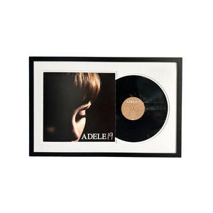 May include: A black framed record album display with a black vinyl record and a photo of a woman's face. The album cover has the text "ADELE 19" printed on it. The vinyl record label has the text "ADELE 19" printed on it.