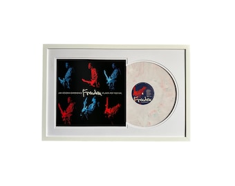 May include: A white framed display with a black and white record album featuring a red and blue image of Jimi Hendrix playing guitar. The album title is "Freedom" and the text "Jimi Hendrix Experience" and "Atlanta Pop Festival" are also visible. The record is displayed next to a white vinyl record with a pink and blue swirl design.