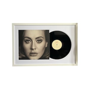 May include: A white framed picture of a woman's face with a black vinyl record in a separate frame. The record label says "25" in red.