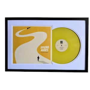 May include: A black framed record display with a white mat. The record is yellow and features a black and white image of a man walking on a winding road with a rocket in the sky. The album cover features the text "Bruno Mars" and "Doo-Wops & Hooligans".