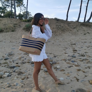 Peut inclure: Une femme en robe blanche à œillets et lunettes de soleil marche sur une plage de sable avec un grand sac fourre-tout rayé.
