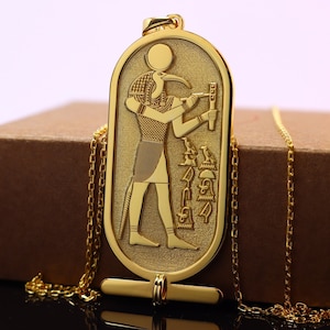 Puede incluir: Collar con colgante dorado con un diseño jeroglífico egipcio grabado. El colgante representa una figura con cabeza de pájaro, sosteniendo un bastón, y está suspendido de una delicada cadena. El collar tiene un cierre de barra en T.