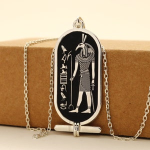 Puede incluir: Un collar de cadena de plata con un colgante negro y plateado que representa a un dios egipcio con cabeza de animal. El colgante es rectangular y tiene jeroglíficos en el lateral.