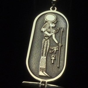 May include: A silver pendant necklace with an oval-shaped charm featuring an Egyptian god with a staff and an ankh symbol. The charm has a textured surface and a gold-colored design.