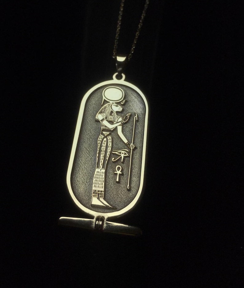 May include: A silver pendant necklace with an oval-shaped charm featuring an Egyptian god with a staff and an ankh symbol. The charm has a textured surface and a gold-colored design.