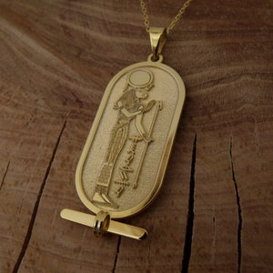 May include: A gold pendant necklace featuring an oval-shaped charm with an embossed image of an Egyptian goddess holding a staff. The charm has a textured surface and a small loop at the top for attaching to a chain.