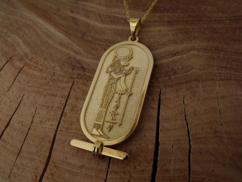 May include: A gold pendant necklace featuring an oval-shaped charm with an embossed image of an Egyptian goddess holding a staff. The charm has a textured surface and a small loop at the top for attaching to a chain.