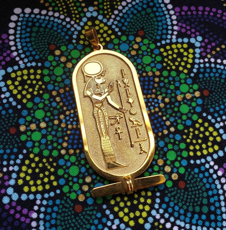 May include: A gold pendant featuring an Egyptian goddess with a sun disc above her head. The pendant is in the shape of a cartouche with hieroglyphs on the sides. The pendant is on a black background with a pattern of small circles.