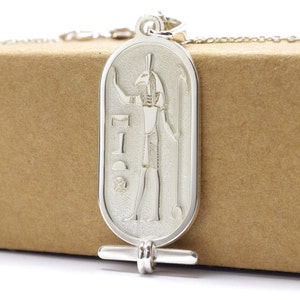 Puede incluir: Pendiente de plata con un dios egipcio con cabeza de halcón, sosteniendo un bastón. El pendiente tiene la forma de un cartucho, un óvalo protector.