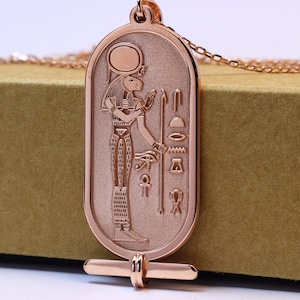 May include: A rose gold pendant necklace featuring an Egyptian goddess with a lion head, holding a staff. The pendant is oval shaped and has a detailed design with hieroglyphics.