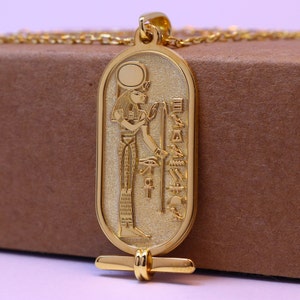 May include: Gold pendant necklace featuring an Egyptian goddess with a sun disk above her head. The pendant is in the shape of a cartouche with hieroglyphs on the sides.
