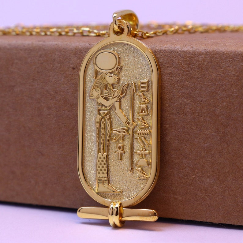 May include: Gold pendant necklace featuring an Egyptian goddess with a sun disk above her head. The pendant is in the shape of a cartouche with hieroglyphs on the sides.