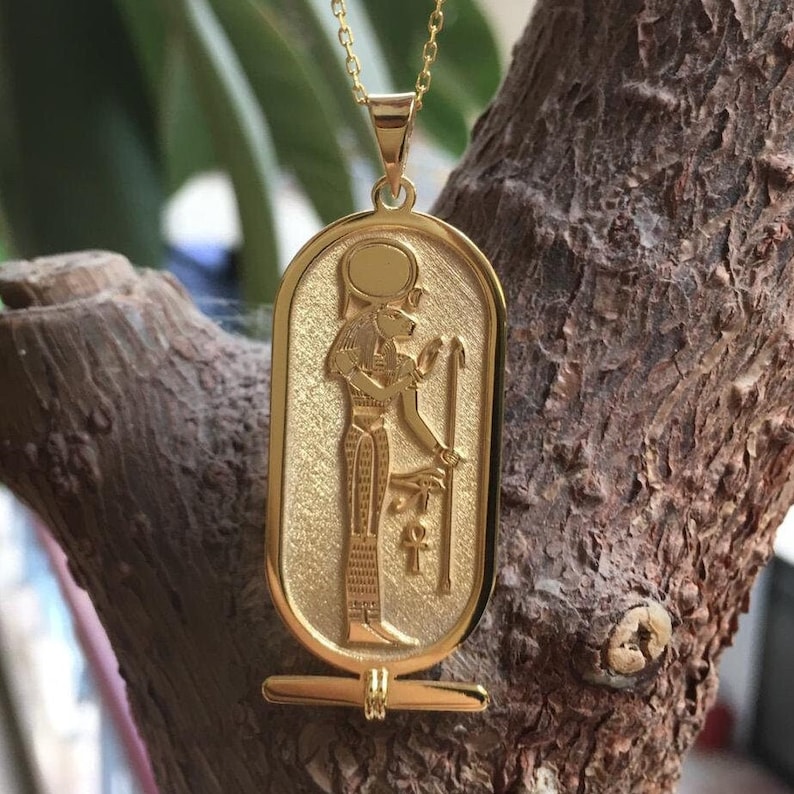 May include: A gold pendant necklace featuring an Egyptian goddess with a lion head, holding a staff and an ankh symbol. The pendant is in the shape of a rectangular cartouche.