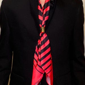 Könnte beinhalten: Ein schwarzer Blazer mit einem rot-schwarz gestreiften Schal, der um den Hals gebunden ist. Der Schal wird mit einem goldenen Ring gehalten.