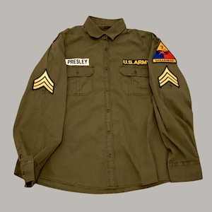 Pode incluir: Camisa militar verde-oliva com dois bolsos no peito, gola e fecho de botões. Apresenta o nome "PRESLEY" numa etiqueta de identificação, um patch "U.S. ARMY" e um patch "SPEARHEAD". Galões dourados nas mangas.
