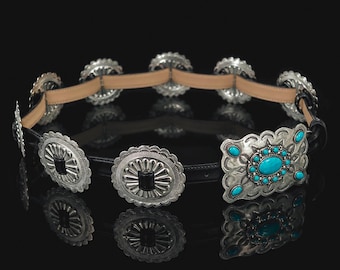 May include: A black leather belt featuring a series of circular silver-toned metal conchos with black center accents and a large rectangular silver-toned buckle adorned with multiple bright turquoise-colored oval stones in a decorative pattern.