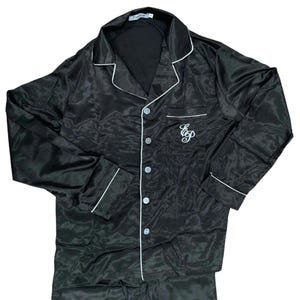 Puede incluir: Camiseta de pijama de satén negro con ribetes blancos y un logotipo con monograma en el pecho. La camiseta tiene un cierre de botones en la parte delantera y mangas largas.