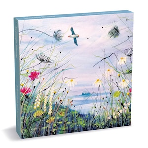 May include: A square box set titled Seascape Blooms containing 8 notecards with envelopes. The box features a coastal illustration with a flying puffin, white daisies, pink flowers, and tall grasses against a blue sea and sky background.