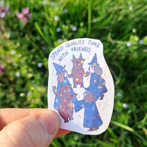 Op de afbeelding: Een holografische sticker met vijf cartoonfiguren in heksenkostuums. De sticker heeft de tekst "Spend quality time with friends". De figuren zijn in tinten blauw en bruin.