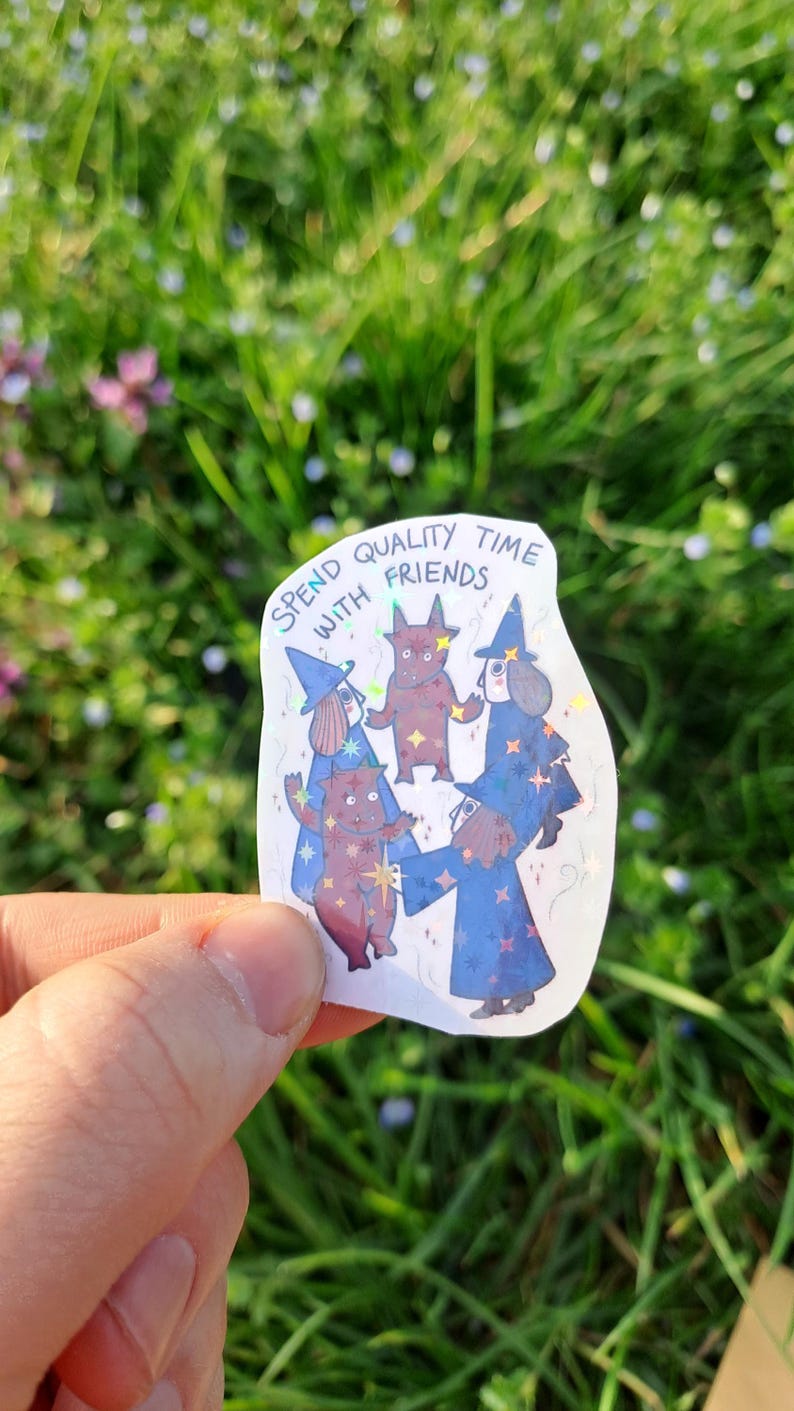 Op de afbeelding: Een holografische sticker met vijf cartoonfiguren in heksenkostuums. De sticker heeft de tekst "Spend quality time with friends". De figuren zijn in tinten blauw en bruin.