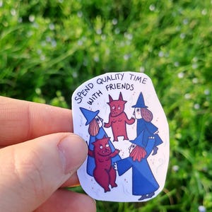 Op de afbeelding: Een witte sticker met de tekst "Spend quality time with friends" en illustraties van striptekeningen van heksen en rode duivels. De sticker wordt tegen een achtergrond van groen gras gehouden.