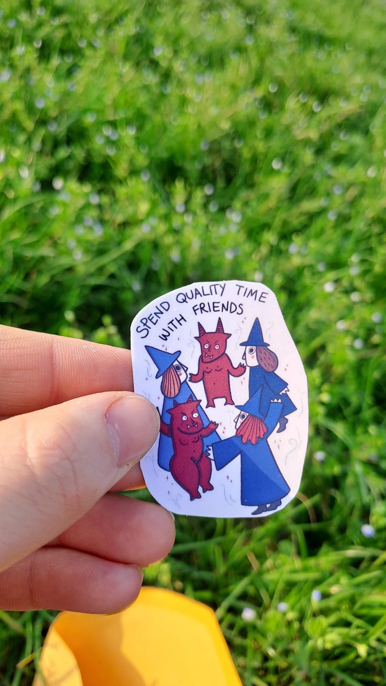 Op de afbeelding: Een witte sticker met de tekst "Spend quality time with friends" en illustraties van striptekeningen van heksen en rode duivels. De sticker wordt tegen een achtergrond van groen gras gehouden.