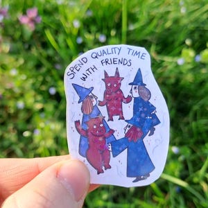 Op de afbeelding: Een sticker met de tekst "SPEND QUALITY TIME WITH FRIENDS" toont cartoonfiguren van heksen en rode wezens. De heksen dragen blauwe gewaden en hoeden. De sticker wordt tegen een achtergrond van groen gras en bloemen gehouden.