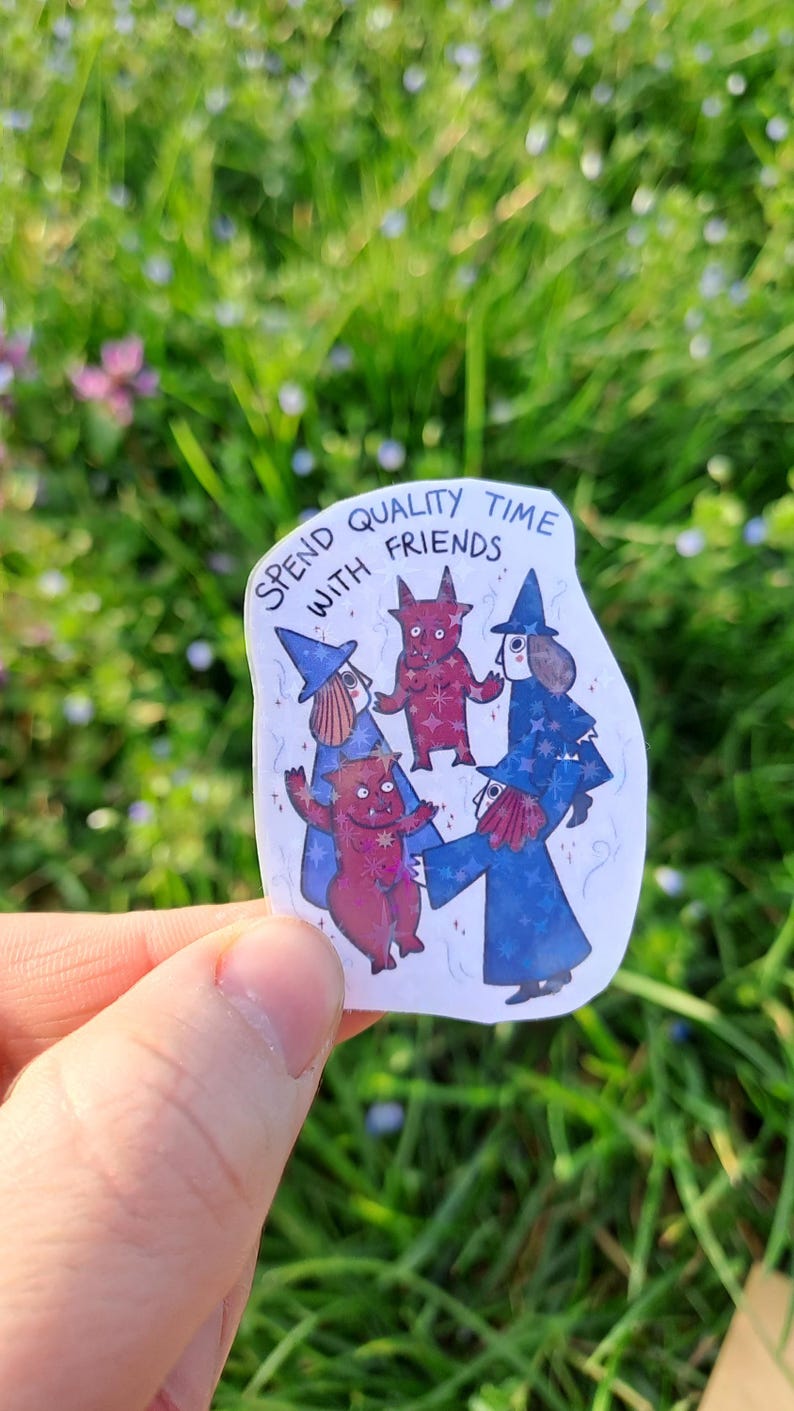 Op de afbeelding: Een sticker met de tekst "SPEND QUALITY TIME WITH FRIENDS" toont cartoonfiguren van heksen en rode wezens. De heksen dragen blauwe gewaden en hoeden. De sticker wordt tegen een achtergrond van groen gras en bloemen gehouden.