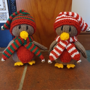Könnte beinhalten: Zwei gestrickte Pinguin-Ornamente mit rot-weiß gestreiften Mützen und Schals. Der Pinguin links hat einen grün-rot gestreiften Schal, während der Pinguin rechts einen weiß-rot gestreiften Schal hat. Beide Pinguine haben gelbe Schnäbel und Füße.