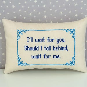 May include: A rectangular off-white throw pillow featuring blue cross-stitch embroidery. The text reads, I'll wait for you. Should I fall behind, wait for me. The lettering is enclosed within a decorative blue cross-stitch border frame.