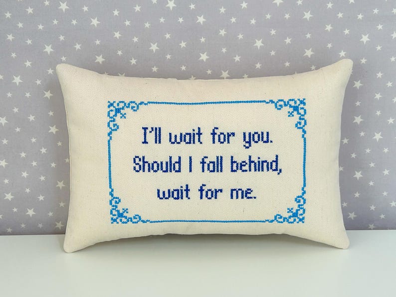 May include: A rectangular off-white throw pillow featuring blue cross-stitch embroidery. The text reads, I'll wait for you. Should I fall behind, wait for me. The lettering is enclosed within a decorative blue cross-stitch border frame.