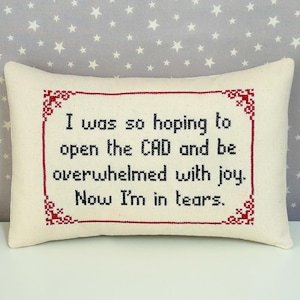 A rectangular pillow made with cream fabric. The text "I was so hoping to open the CAD and be overwhelmed with joy. Now I'm in tears." is cross stitch embroidered on the pillow in lowercase using black thread. The pillow has a maroon cross stitched border with a classic design cross stitched on each corner of the border.