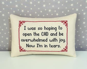 May include: A rectangular off-white throw pillow featuring black cross-stitched text that reads: I was so hoping to open the CAD and be overwhelmed with joy. Now I'm in tears. The text is enclosed in a decorative red cross-stitched border.