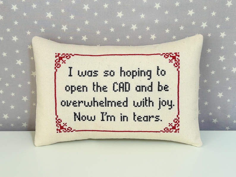 A rectangular pillow made with cream fabric. The text "I was so hoping to open the CAD and be overwhelmed with joy. Now I'm in tears." is cross stitch embroidered on the pillow in lowercase using black thread. The pillow has a maroon cross stitched border with a classic design cross stitched on each corner of the border.