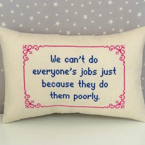 May include: A rectangular off-white throw pillow featuring a cross-stitched design. The center text reads, "We can't do everyone's jobs just because they do them poorly." The text is in blue, surrounded by a decorative pink cross-stitched border.
