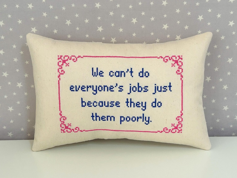 May include: A rectangular off-white throw pillow featuring a cross-stitched design. The center text reads, "We can't do everyone's jobs just because they do them poorly." The text is in blue, surrounded by a decorative pink cross-stitched border.