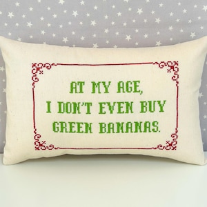 May include: A rectangular off-white throw pillow featuring a cross-stitched design. The text reads, "AT MY AGE, I DON'T EVEN BUY GREEN BANANAS." in green, enclosed within a decorative red cross-stitched border with ornate corner details.