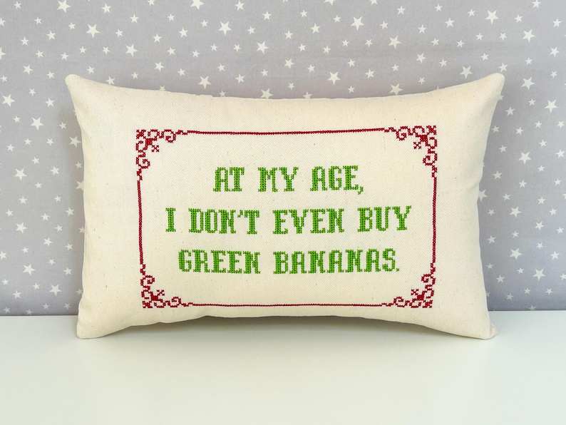 May include: A rectangular off-white throw pillow featuring a cross-stitched design. The text reads, "AT MY AGE, I DON'T EVEN BUY GREEN BANANAS." in green, enclosed within a decorative red cross-stitched border with ornate corner details.