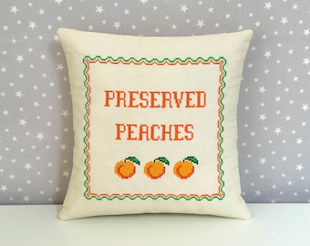 May include: A square off-white throw pillow featuring a cross-stitched design. The center displays the orange text PRESERVED PEACHES above three small orange peaches with green leaves, all enclosed within a decorative green and orange border.
