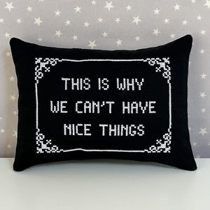 May include: A rectangular black throw pillow featuring white cross-stitch embroidery. The center text reads "THIS IS WHY WE CAN'T HAVE NICE THINGS" enclosed within a decorative white cross-stitch border with ornate corner details.