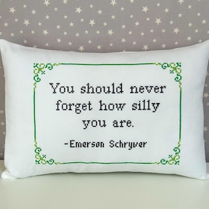 May include: A rectangular white throw pillow featuring a cross-stitched design in black and green. The text reads, "You should never forget how silly you are. -Emerson Schryver," enclosed within a decorative green cross-stitched border.
