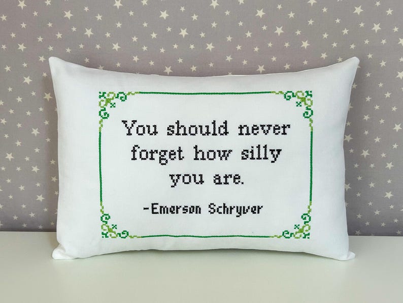 May include: A rectangular white throw pillow featuring a cross-stitched design in black and green. The text reads, "You should never forget how silly you are. -Emerson Schryver," enclosed within a decorative green cross-stitched border.