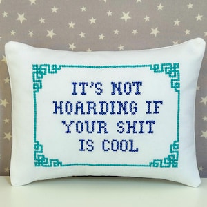 May include: A rectangular white throw cushion featuring a cross-stitched border in teal and dark blue text that reads, IT'S NOT HOARDING IF YOUR SHIT IS COOL. The embroidery is centred on the cushion, which has a smooth fabric finish and clean edges.
