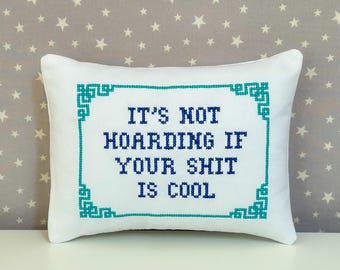 May include: A rectangular white throw pillow featuring a cross-stitched border in teal and dark blue text that reads, IT'S NOT HOARDING IF YOUR SHIT IS COOL. The embroidery is centered on the pillow, which has a smooth fabric finish and clean edges.