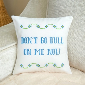May include: A square white throw pillow featuring blue cross-stitch lettering that reads "DON'T GO DULL ON ME NOW." The text is framed by two horizontal rows of green and blue floral cross-stitch patterns at the top and bottom of the pillow.